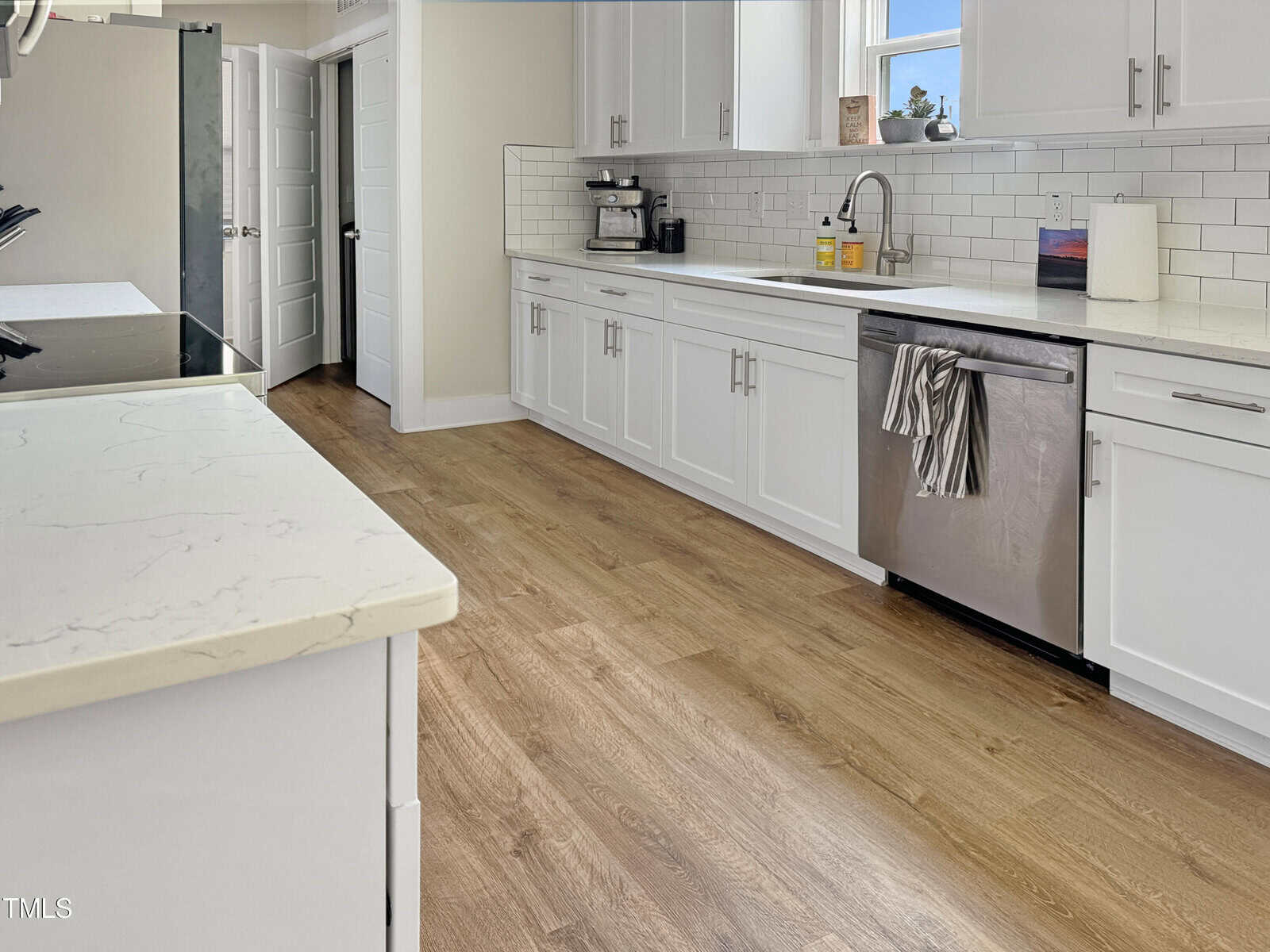 Modern kitchen with white cabinets, stainless steel appliances, subway tile backsplash, a window above the sink, and light wood flooring. A towel hangs on the dishwasher handle, and soap bottles sit by the sink.
