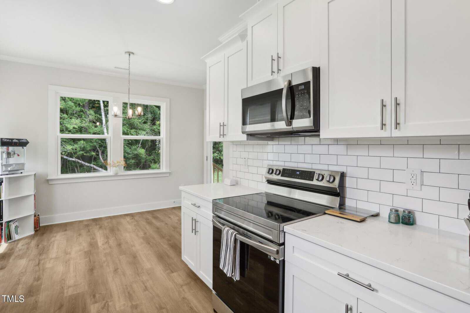 A modern kitchen with white cabinets, stainless steel appliances, subway tile backsplash, wood flooring, quartz countertops, and a windowed dining area with a chandelier.