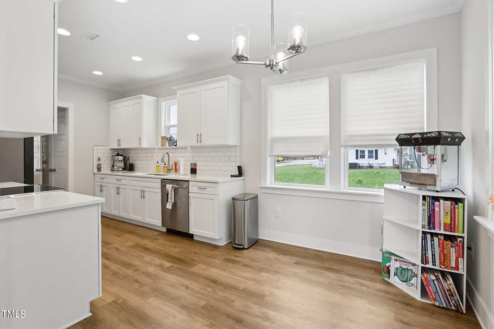 Modern kitchen with white cabinets, stainless steel appliances, wood flooring, and large windows. A bookshelf with games and a popcorn maker sits near the windows. Countertops are clear except for some small appliances and a dish towel.