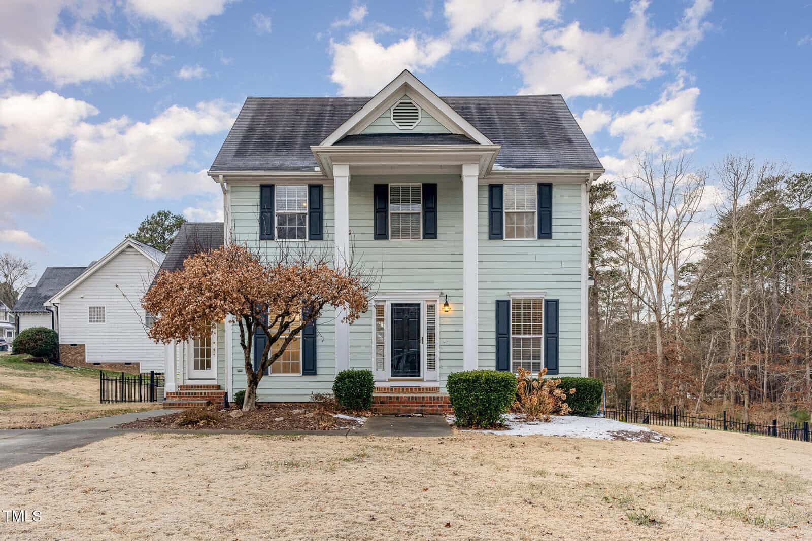 A two-story house with light blue siding and dark shutters. It features a prominent front porch with white columns. The yard has a tree with no leaves and brown grass. A driveway is visible on the left. The background includes trees and a partly cloudy sky.