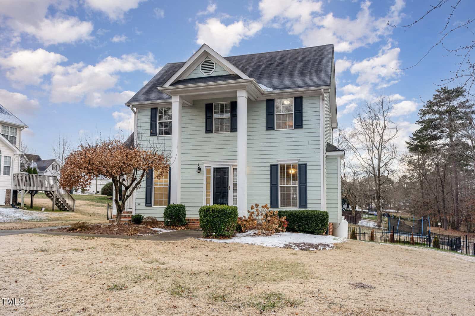 A two-story light blue house with white trim and black shutters is shown. The front features a gable and a small porch with columns. There's a large tree on the left, a fenced backyard, and a patchy lawn with some snow. It's a clear day with scattered clouds.