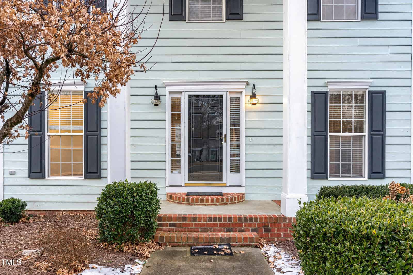 A front exterior view of a house with light blue siding and white trim. There is a glass front door with brick steps leading up to it. Two black shutters frame each window on either side of the door. A leafless tree and small shrubs are in the foreground.