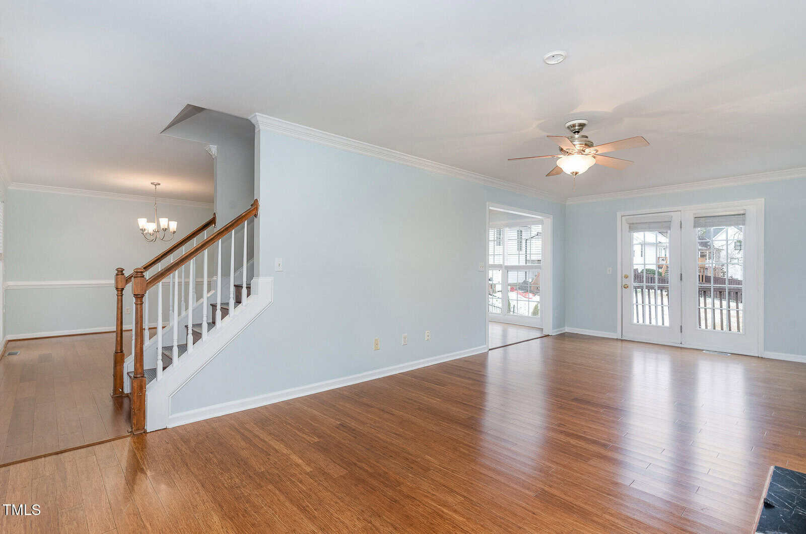 A spacious, empty living room with light blue walls and hardwood flooring. A ceiling fan is mounted above. Wooden stairs with white balusters lead to the upper floor. French doors open to a deck, and a chandelier hangs in the adjacent dining area.