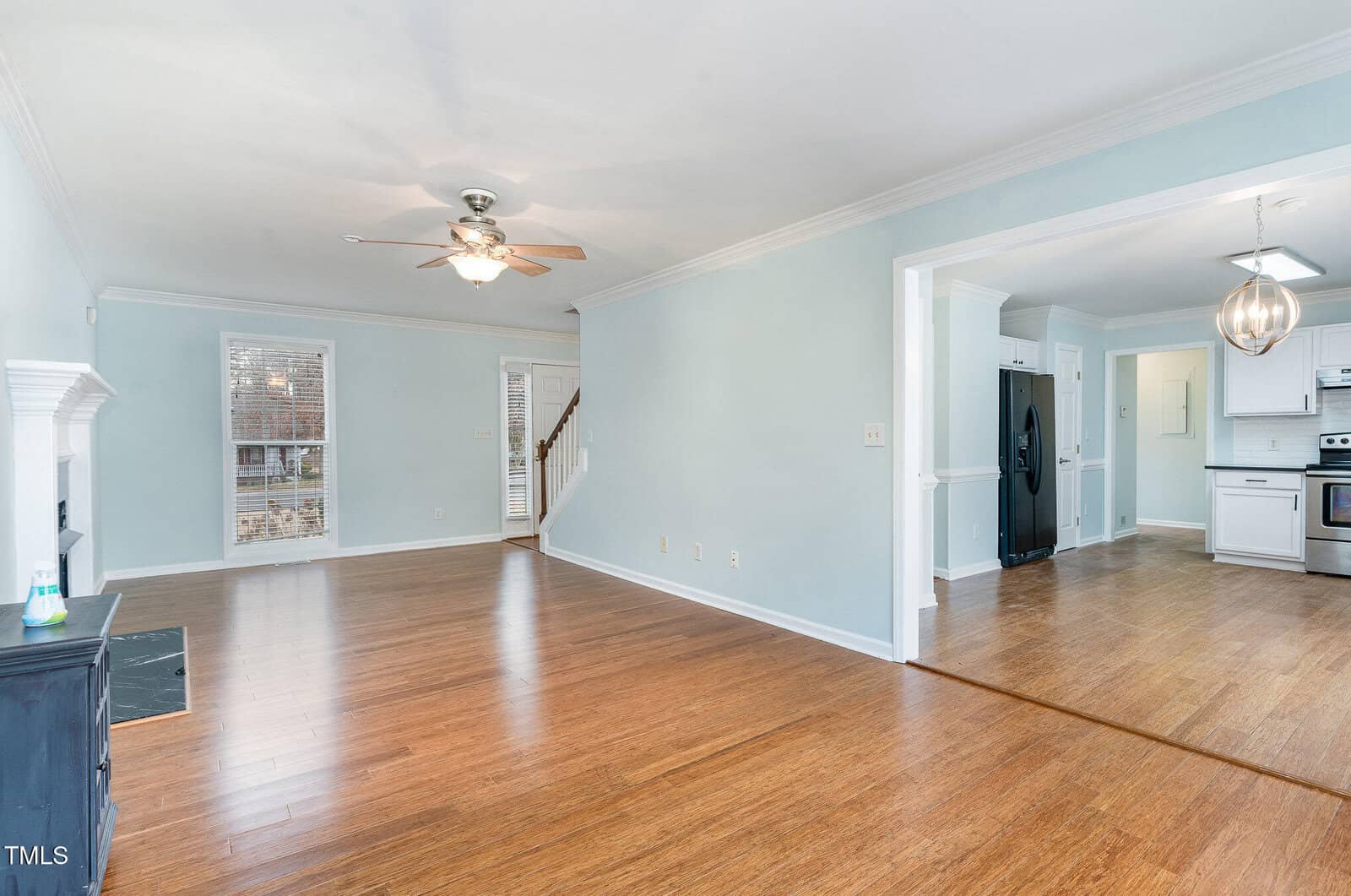A spacious, empty room with light blue walls and wood flooring. The room features a ceiling fan and large windows. There is an adjacent kitchen area with white cabinets and a black refrigerator visible in the background.