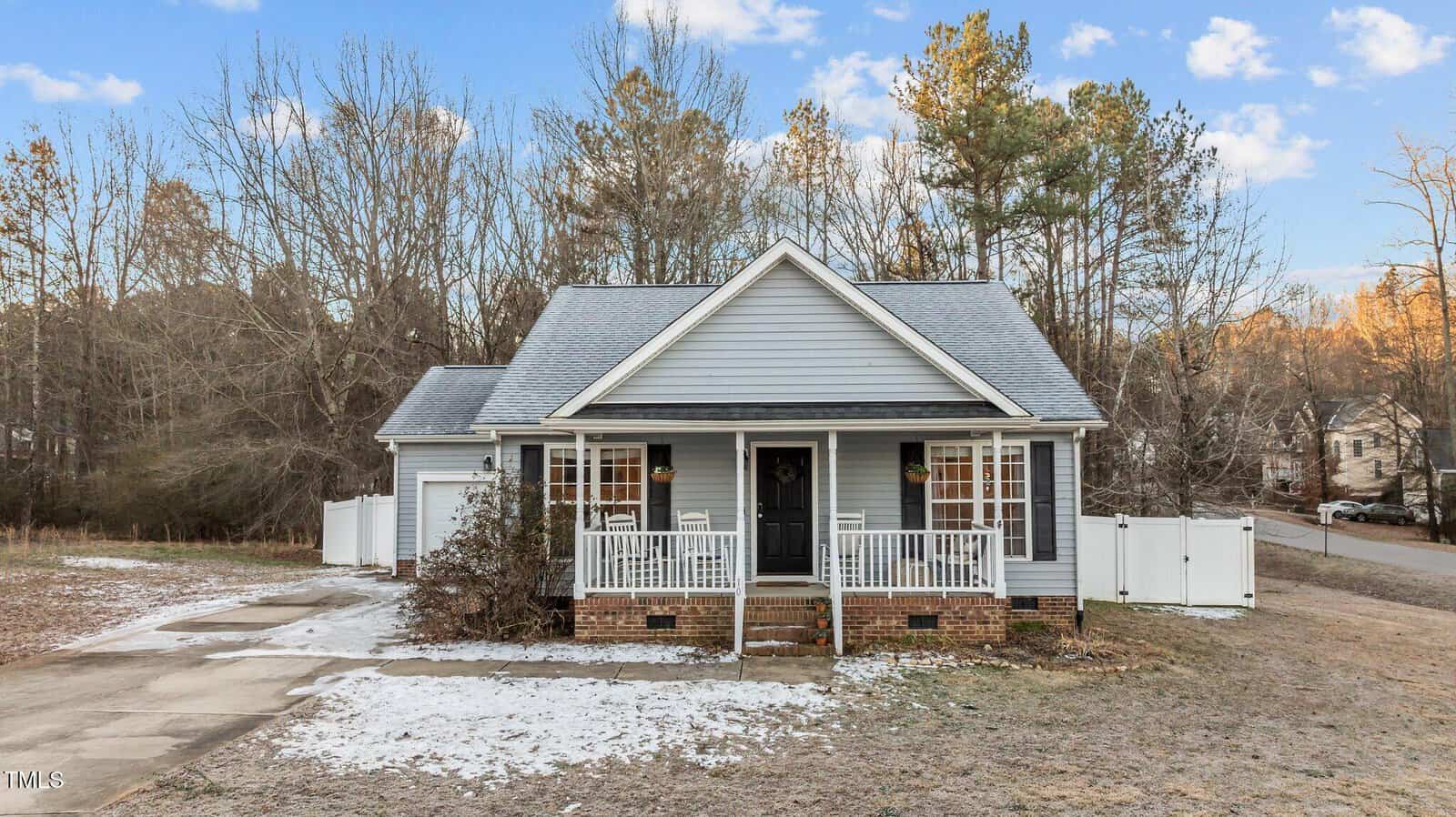 A small, single-story gray house with white trim and a front porch sits on a lot with a partial driveway and light patches of snow on the ground. There are trees and a white fence in the background.