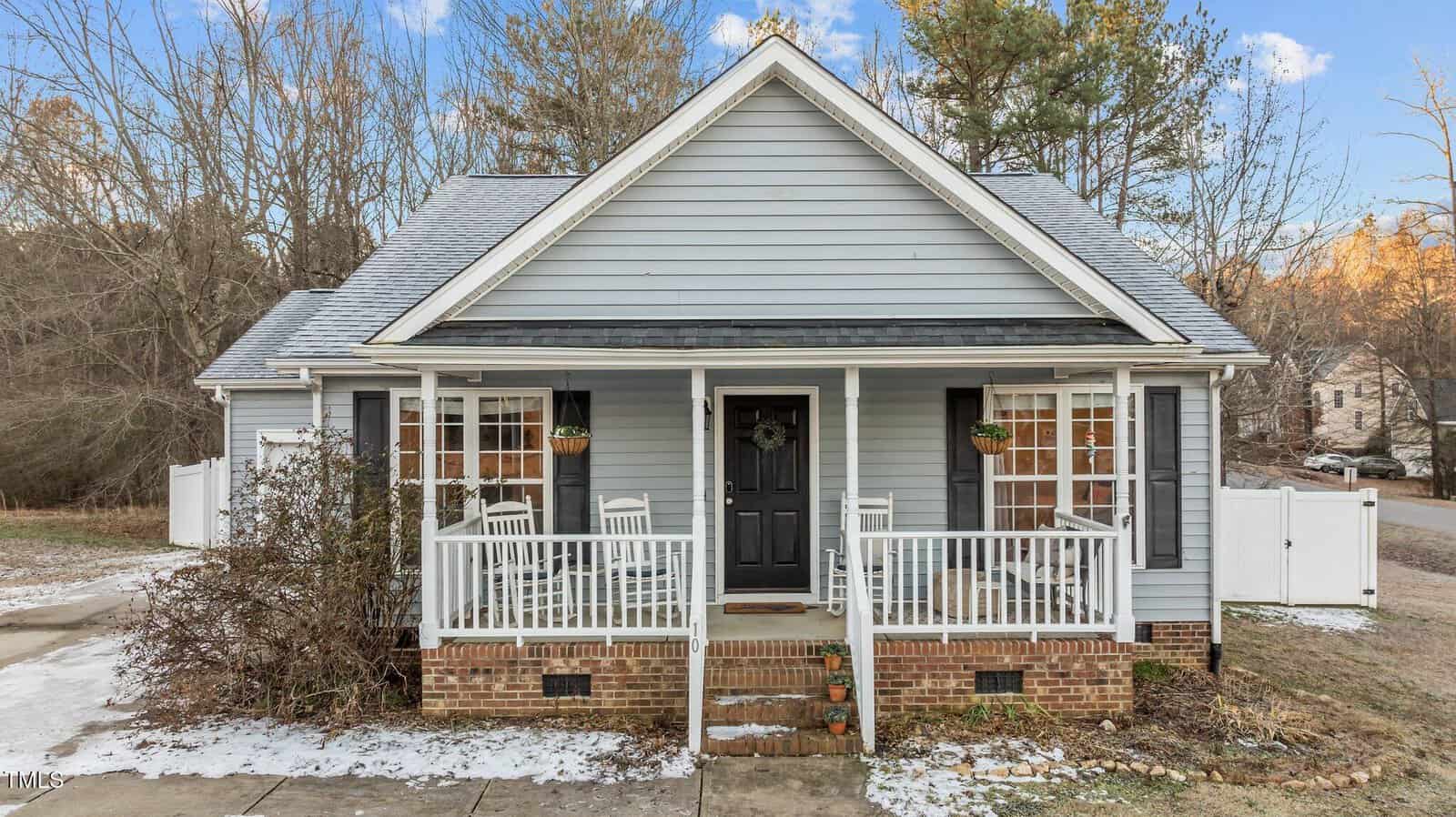 A small, light gray house with white trim, black shutters, and a black front door. The front porch has white railings, steps, rocking chairs, and potted plants. There is a brick foundation and a light dusting of snow on the ground.