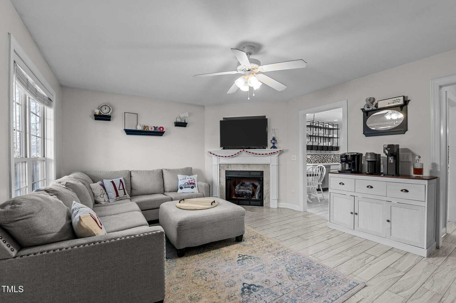 A gray sectional sofa, round ottoman, rug, and wall-mounted TV above a fireplace are in a living room with wood-look flooring. A sideboard with a coffee maker sits by a doorway to a dining area with a patterned wall.