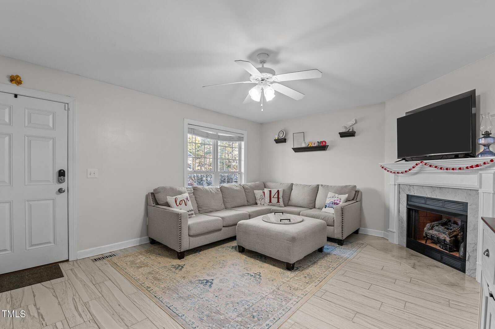A living room with a light gray sectional sofa, ottoman, flat-screen TV above a fireplace, bay window, ceiling fan, light rug, and shelves with decor. The space is decorated in neutral tones with simple accents.