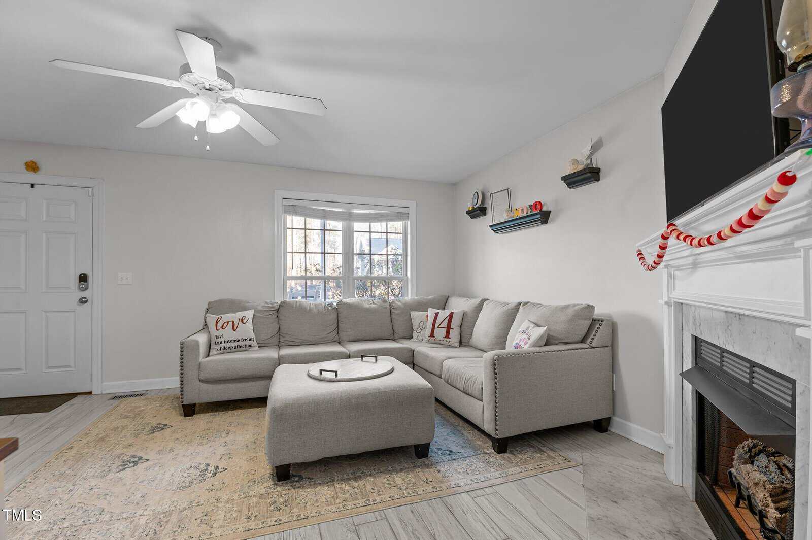 A living room with a gray sectional sofa, decorative pillows, a round ottoman, a fireplace with a striped garland, a ceiling fan, wall shelves, and a window letting in natural light.