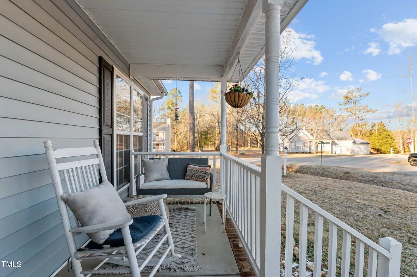 A view of a front porch with a white rocking chair, a patterned rug, a bench with cushions, a hanging planter, and a white railing. The porch overlooks a residential street and trees under a blue sky.