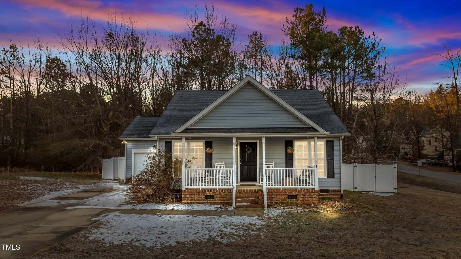 A small, single-story house with gray siding and a dark roof is pictured at dusk. The front porch is lit, and patches of snow cover the ground and driveway. Trees and a white fence are in the background.