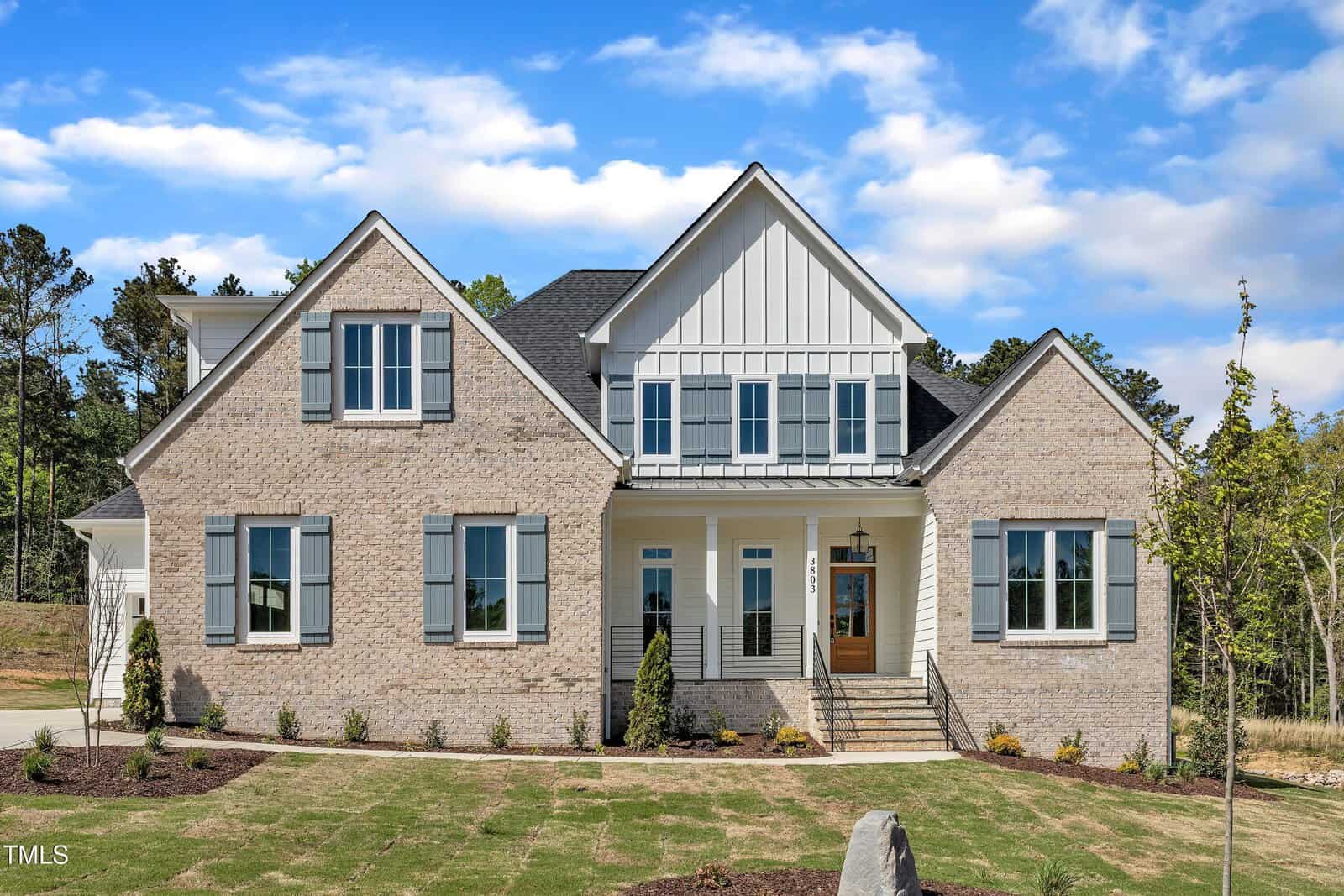 A two-story suburban house at 3803 Bennett Grove Drive, Wake Forest, features a beige brick exterior, blue shutters, a covered front porch, and a neatly landscaped lawn set against trees and a partly cloudy sky.