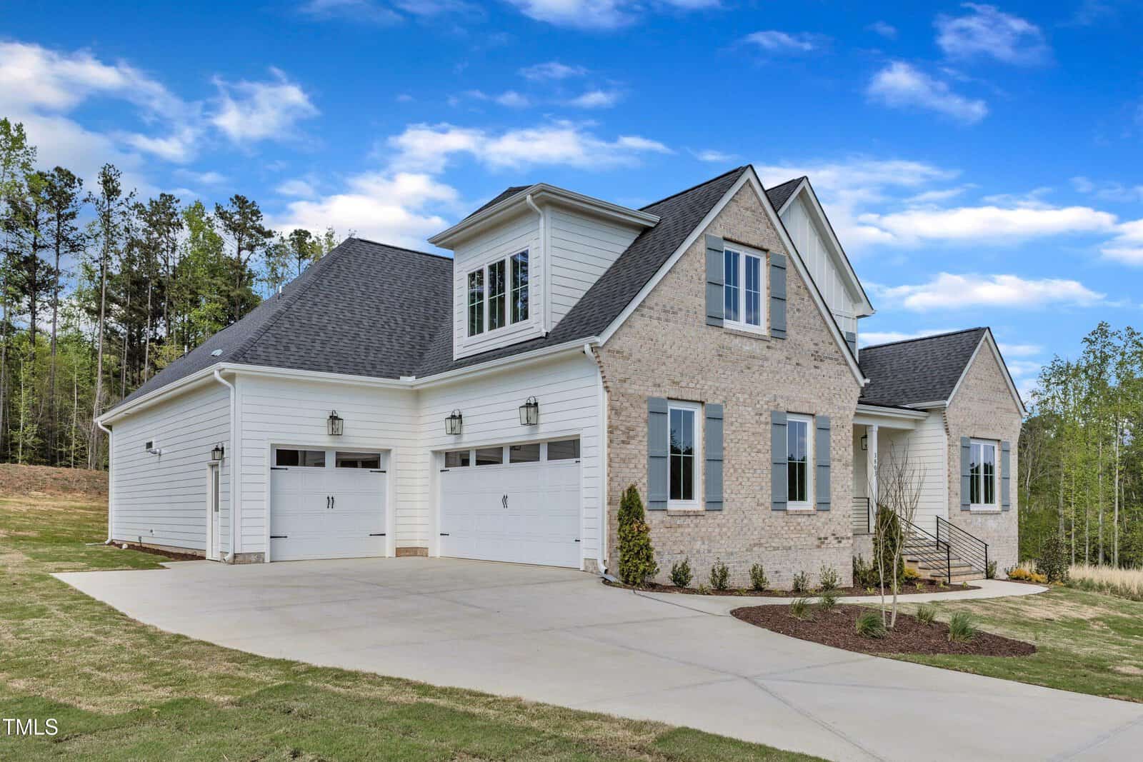 A two-story house with light brick and white siding features three garage doors, a sloped driveway, and a small landscaped front yard, set against a backdrop of trees and a partly cloudy sky.