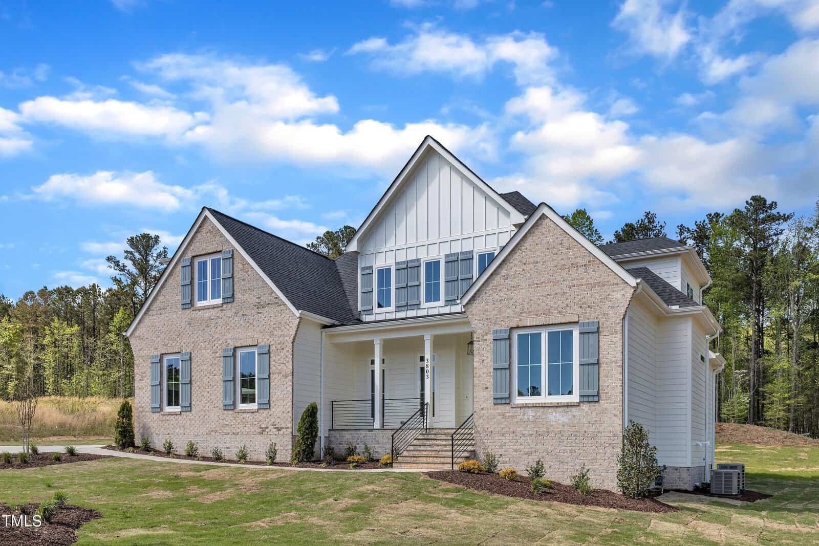 A two-story house with light brick and white siding, black roof shingles, large front windows, and a covered entry porch. The lawn is partially landscaped, with trees and a blue sky in the background.