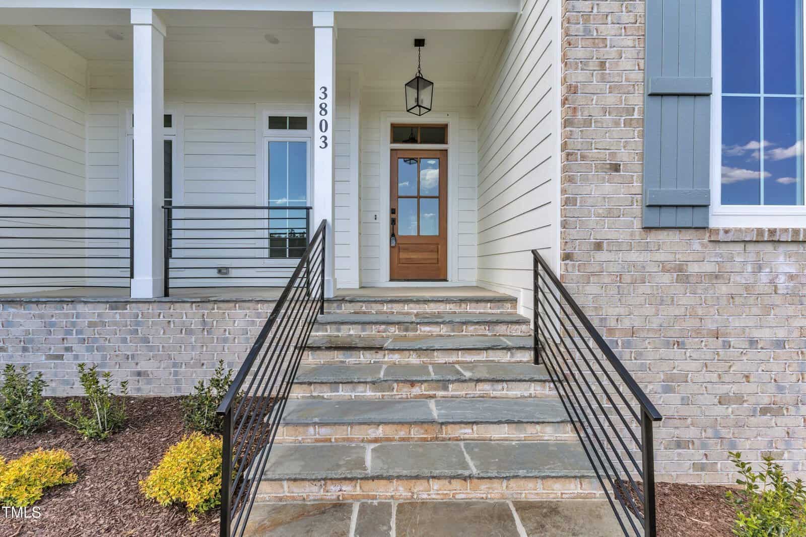 A stone walkway with black metal railings leads up to a wooden front door with glass panels. The entryway features white siding, brick accents, a hanging lantern, and the house number 3803. Shrubs are planted along the path.
