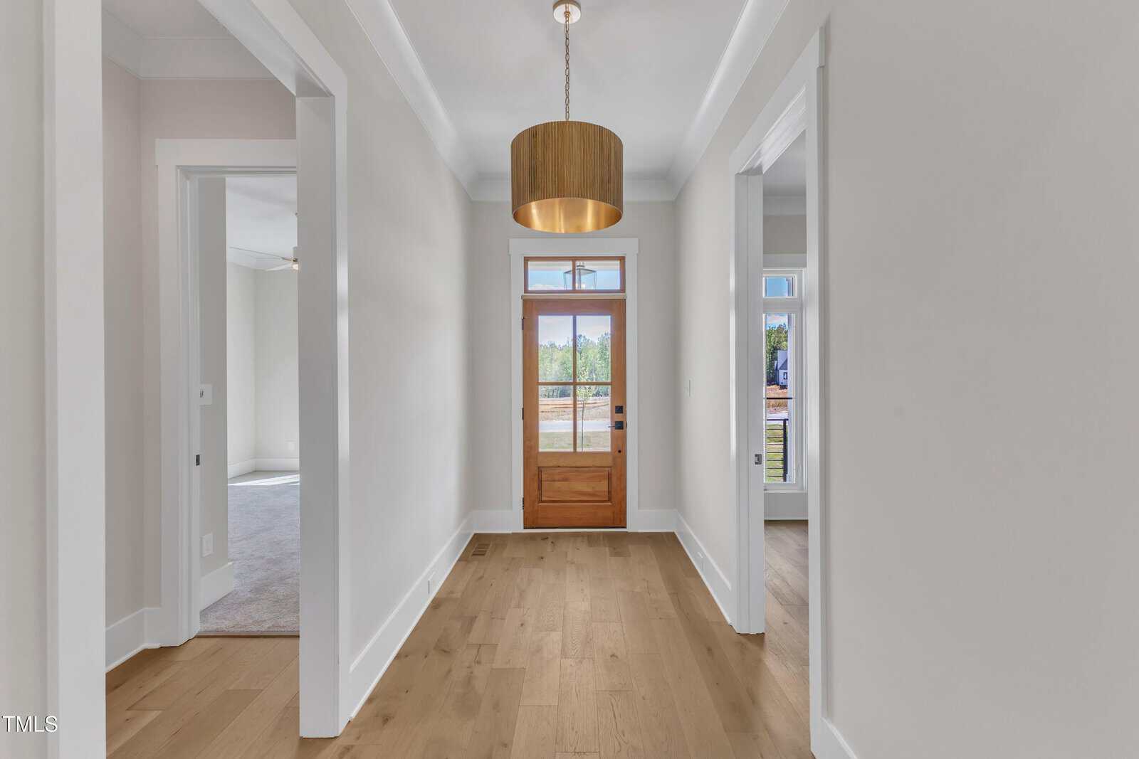A hallway with light-colored walls and wood flooring leads to a wooden front door with glass panels. There are doorways to rooms on both sides and a wooden pendant light hanging from the ceiling.