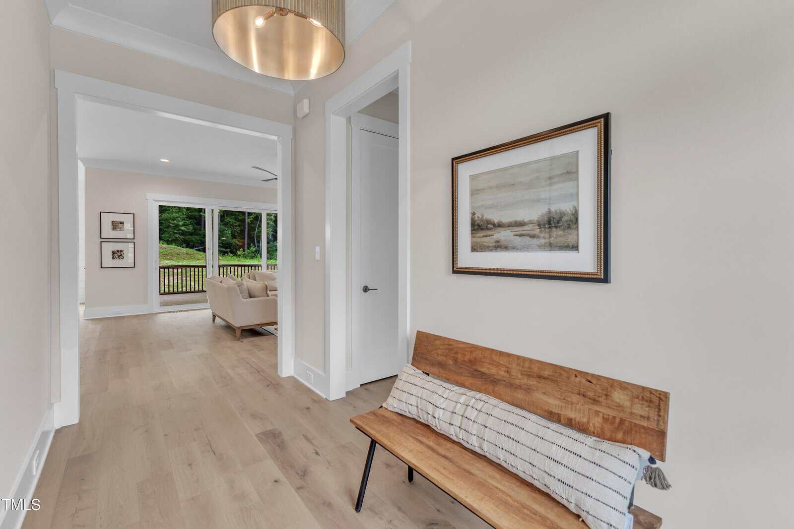 A hallway with light wood flooring, a wooden bench with a striped cushion, a framed landscape picture on the wall, and a modern ceiling light; a living area with sofas is visible in the background.