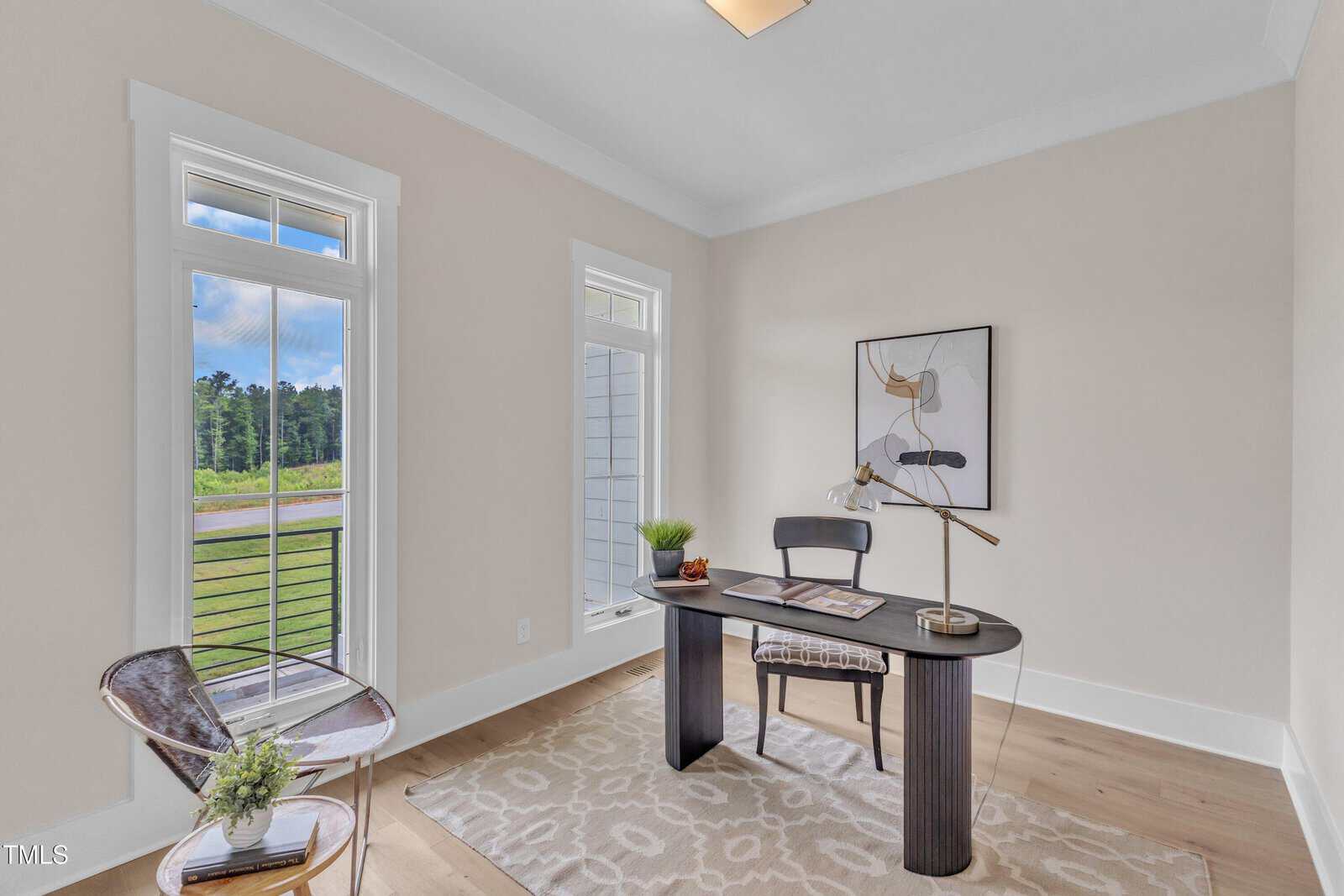 A home office with a modern black desk, chair, desk lamp, and decorative items. There are two tall windows, light-colored walls, wood flooring, a geometric rug, and abstract wall art. A clear chair sits in the corner.