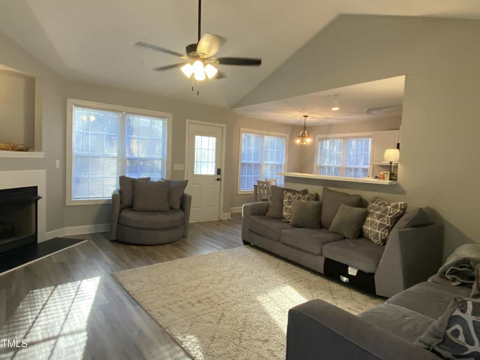 A living room with light gray walls, wood flooring, a ceiling fan, two gray armchairs, a gray sofa with patterned pillows, a white rug, and large windows letting in natural light. Dining area is visible in the back.