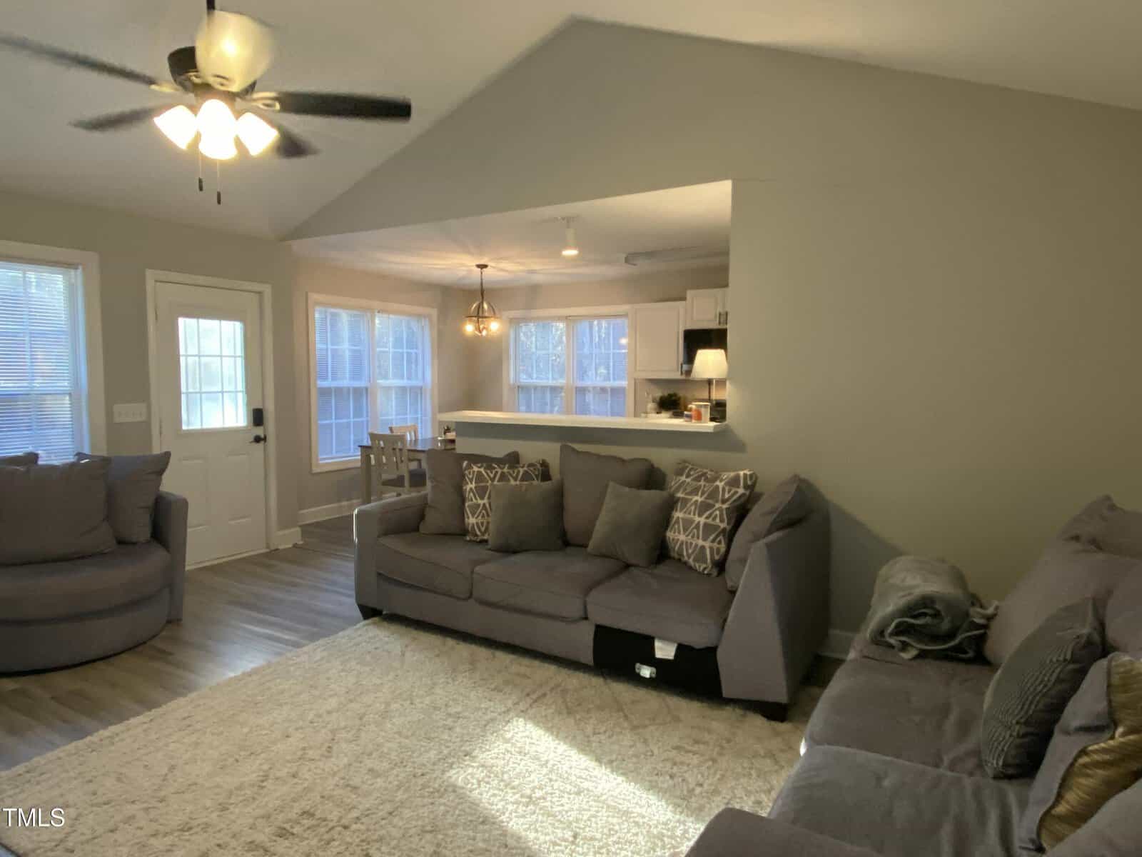 Living room with gray sofas and patterned pillows, round chair, white rug, and ceiling fan. Dining area and kitchen with modern lighting and white cabinets are visible in the background. Neutral walls and wood flooring throughout.