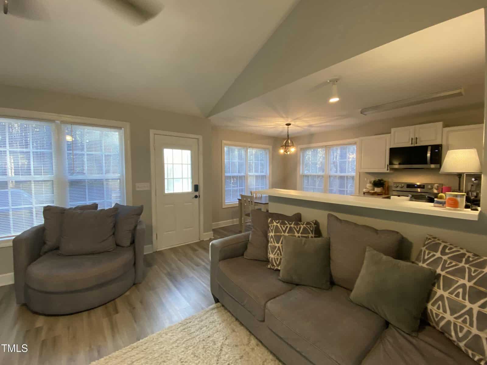 A cozy living room with gray sofas, patterned pillows, a round armchair, and large windows letting in natural light. A small kitchen with white cabinets is visible behind a half wall.