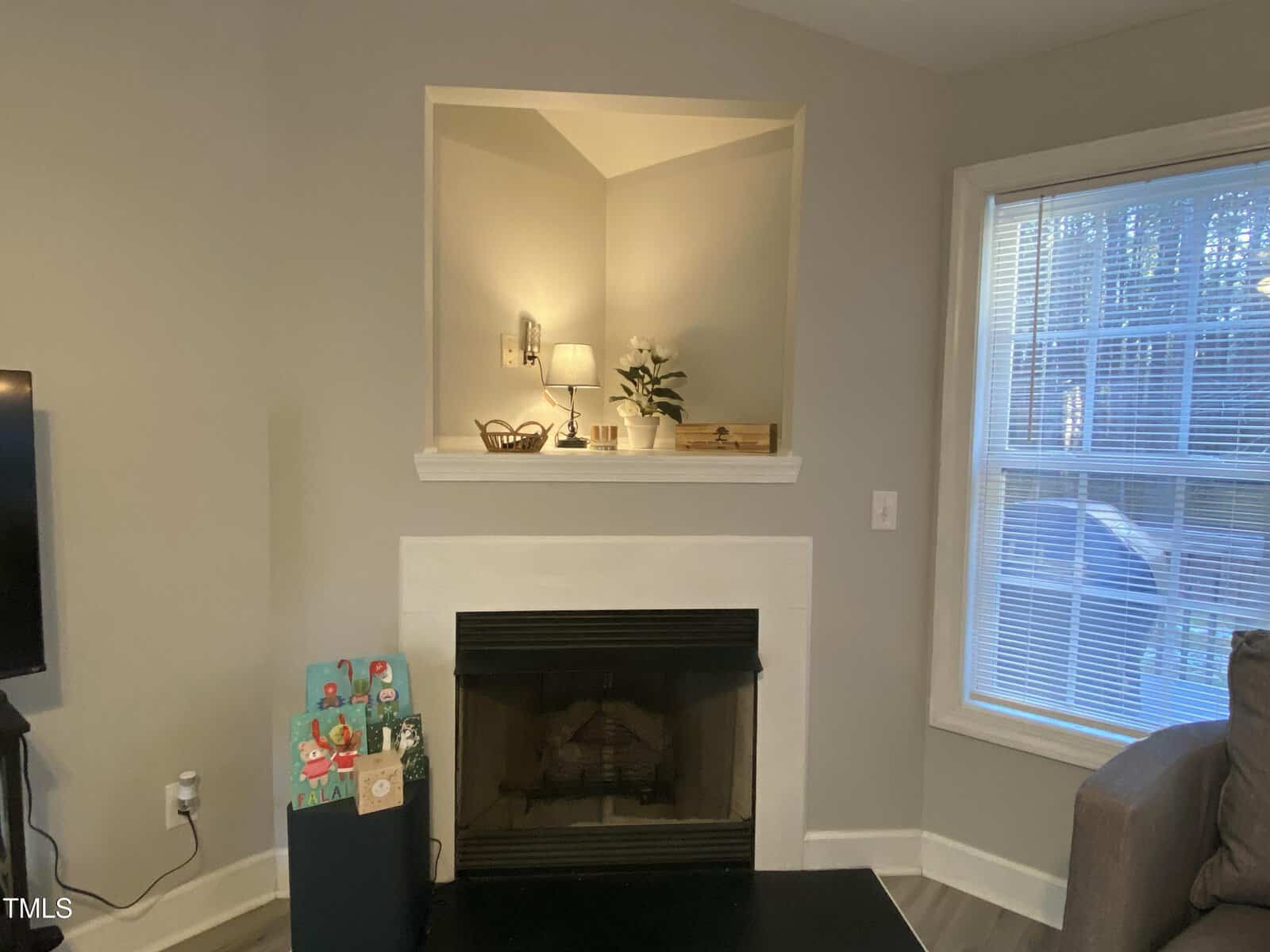 A living room with a fireplace below a recessed shelf holding a lamp, a vase with flowers, and decorative items. To the left, there is a side table with greeting cards, and to the right, a window with closed blinds.