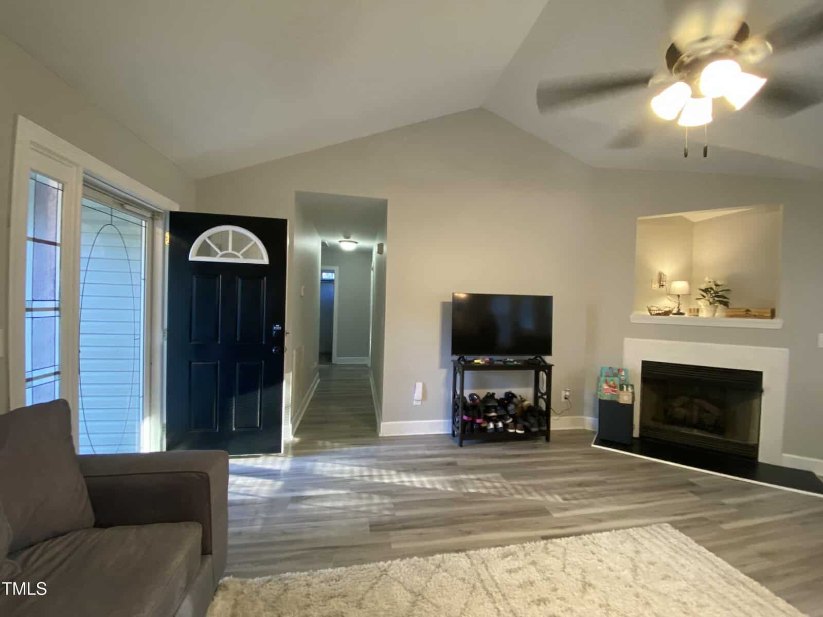 A living room with gray walls, wood flooring, a brown couch, open black front door, TV on a stand, fireplace with decorations, ceiling fan, and light coming through windows on the left.