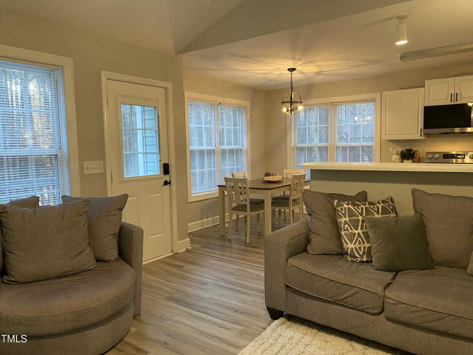 A cozy living room with gray sofas and patterned pillows, next to a dining area with a wooden table and four chairs. Large windows let in natural light. The floor is wood, and the walls are light-colored.