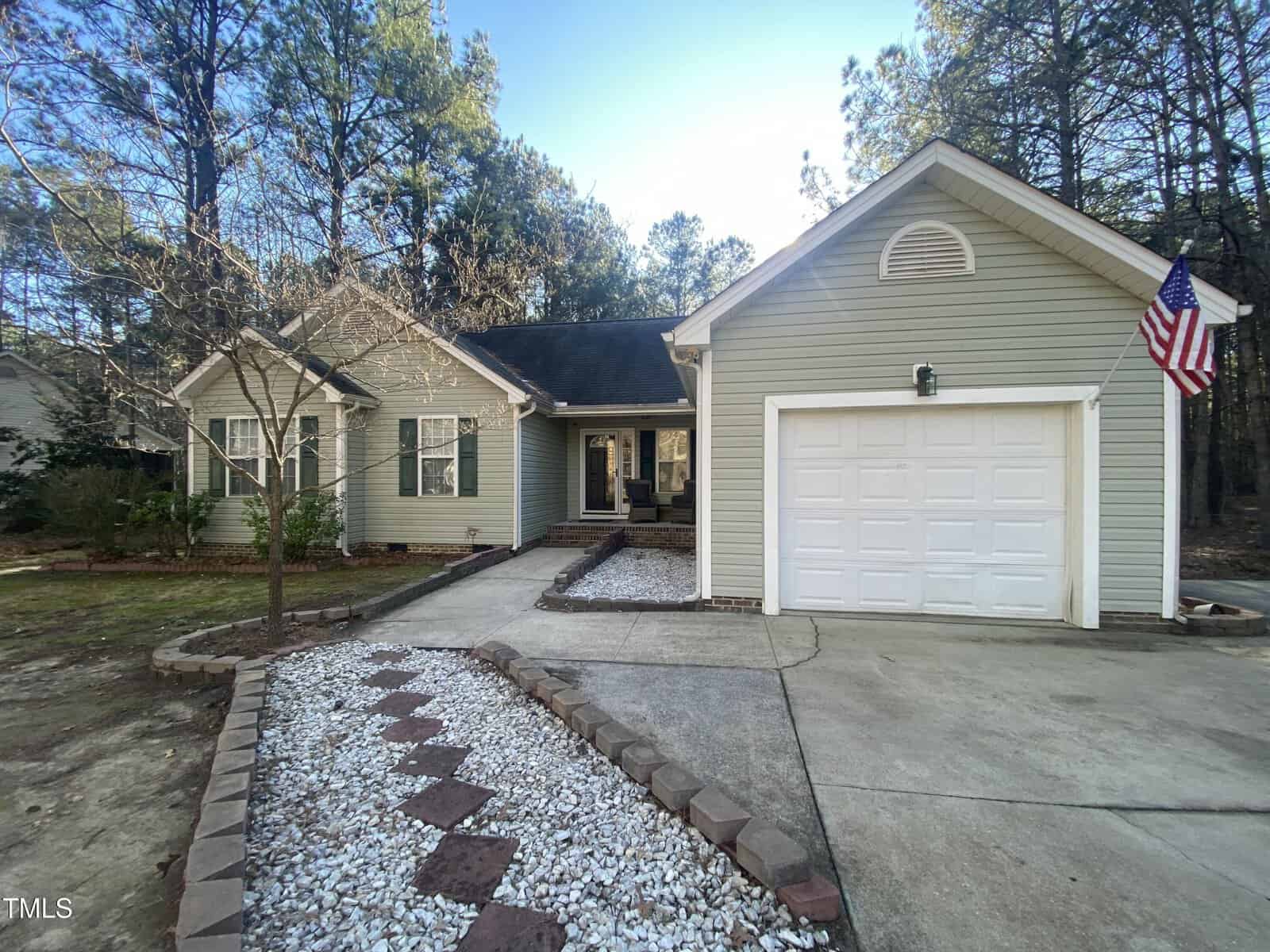 A single-story house with light green siding, white trim, and an attached single-car garage. A small American flag is mounted near the garage. A stone and gravel path leads to the front door, and trees are in the background.