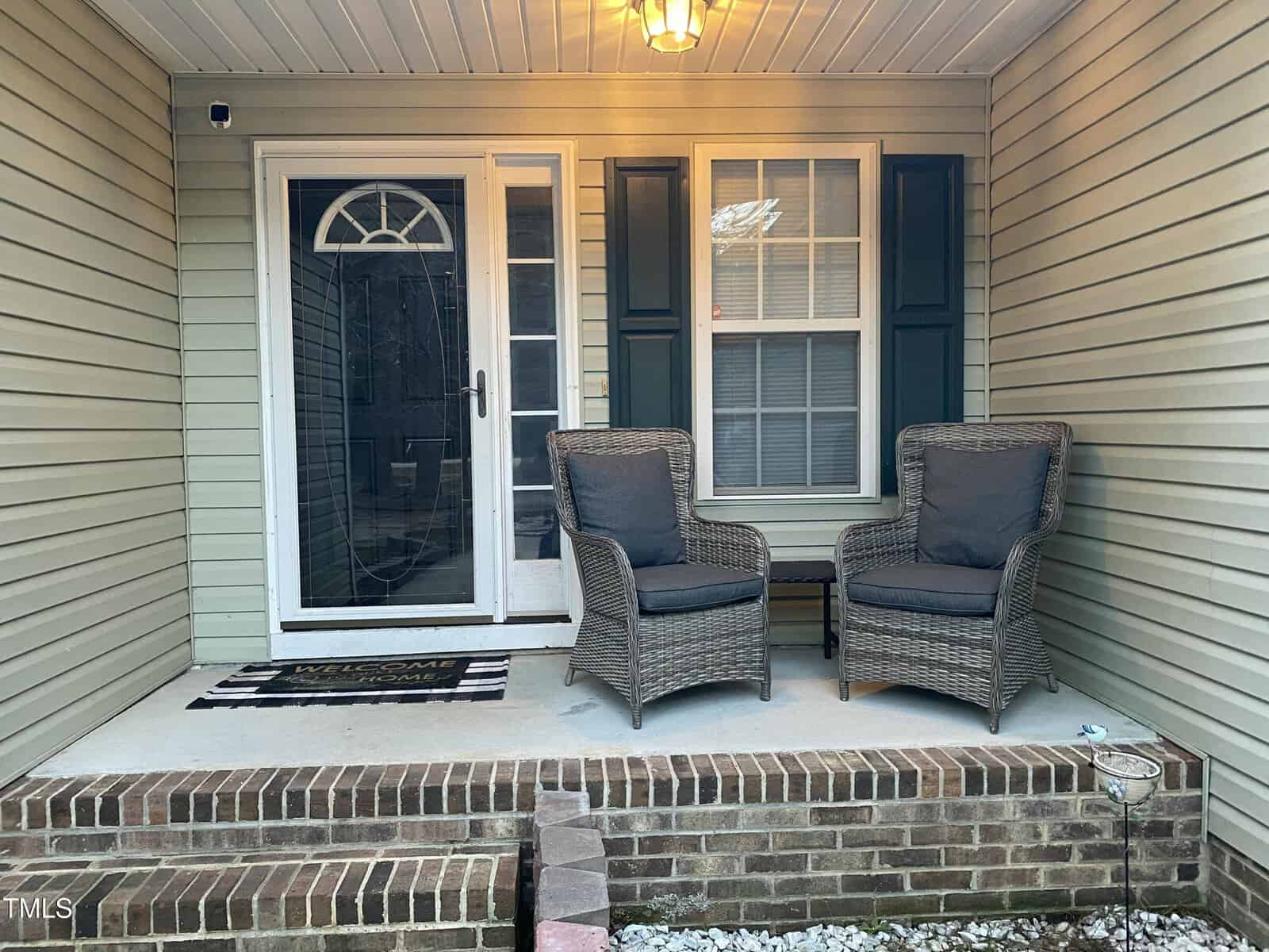 A small covered porch with two wicker chairs with dark cushions, a window with shutters, a glass storm door, a striped doormat, and brick steps leading up to the entrance.