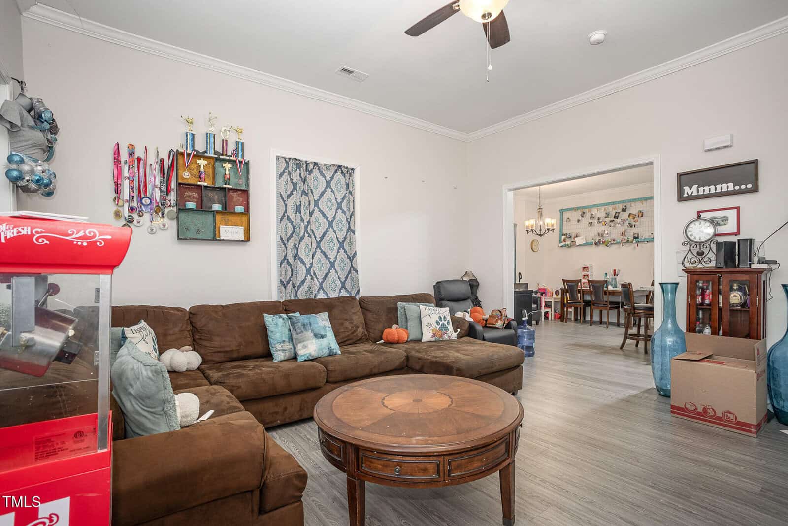 A living room with a brown sectional sofa, a round wooden coffee table, and a red popcorn machine. The walls are decorated with ribbons and trophies. Pumpkins are scattered on the sofa. A ceiling fan hangs above, and an adjoining room is visible.