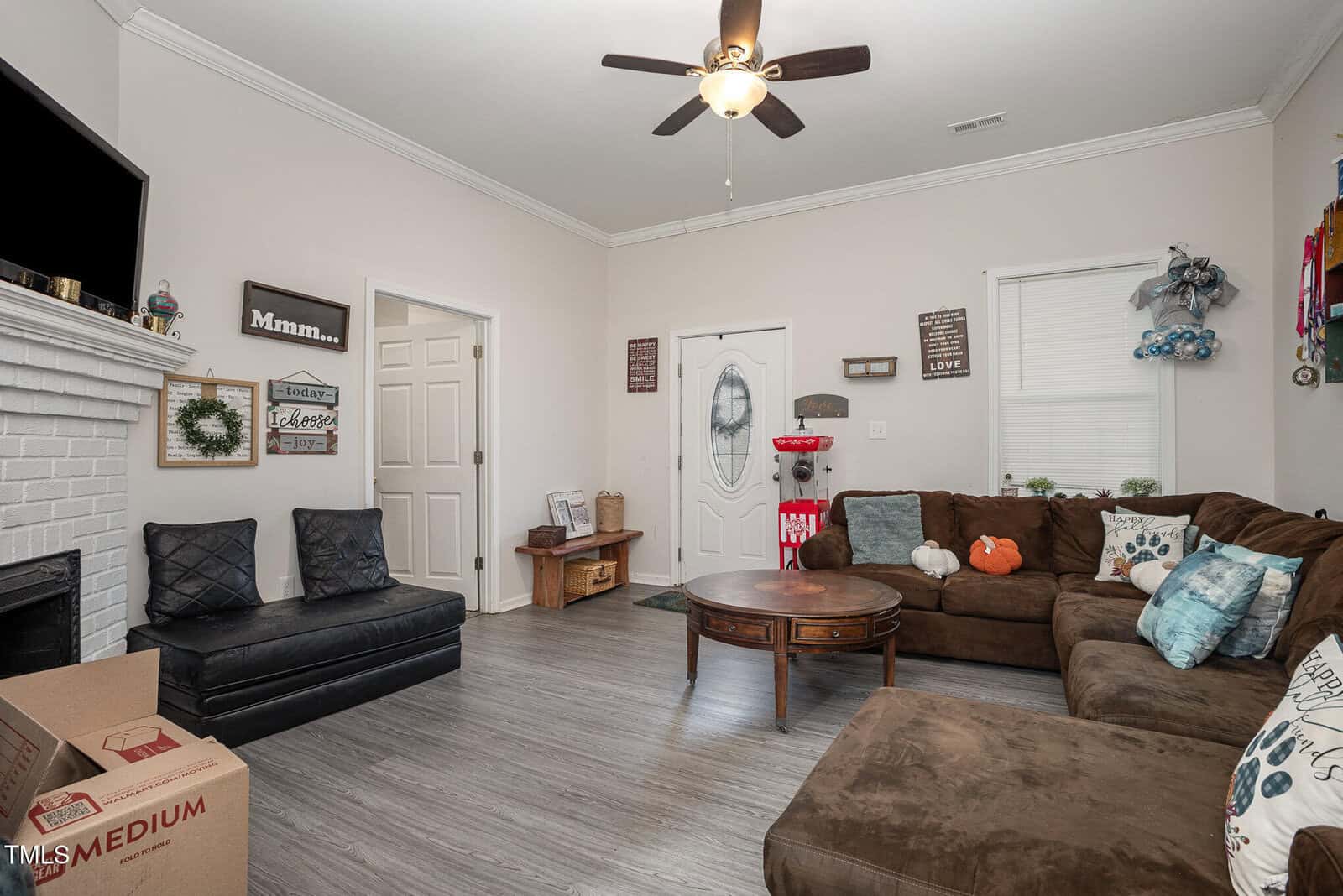 A living room with a brown sectional sofa, a round wooden coffee table, and a wall-mounted TV. Decorative pillows are on the sofa, and various wall art pieces are displayed. Ceiling fan with light fixture is on, and a cardboard box sits on the floor.