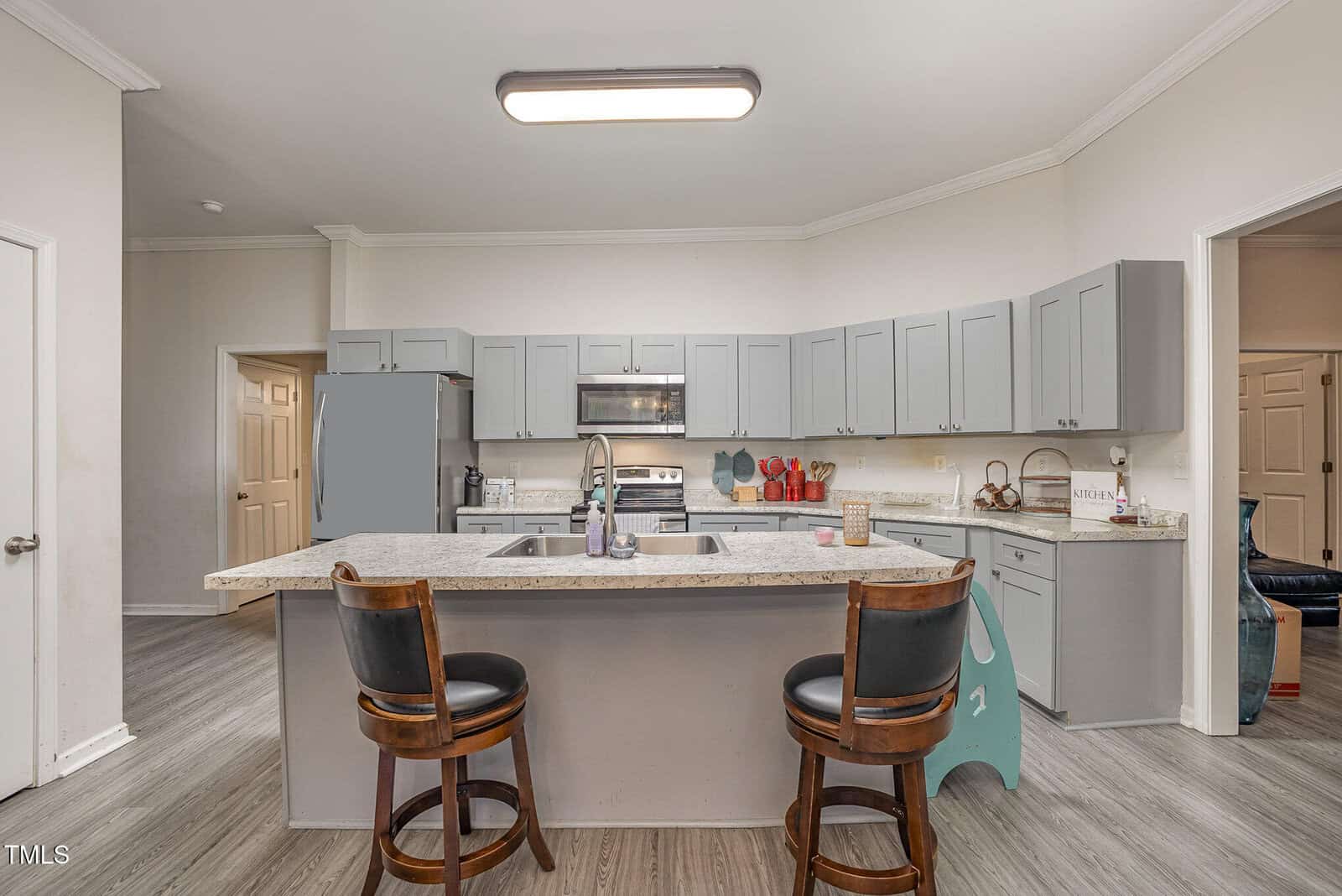 A modern kitchen with gray cabinets, a large island with a sink, two wooden bar stools, and stainless steel appliances. The room features light-colored flooring and a ceiling light fixture. A door leads to another room on the right.