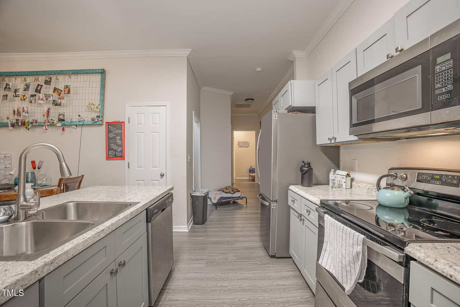 A modern kitchen with light gray cabinets and a stainless steel refrigerator. A black stove with a teal kettle sits near a gray microwave. A kitchen island has a double sink; a corkboard with photos and a chalkboard are on the opposite wall.