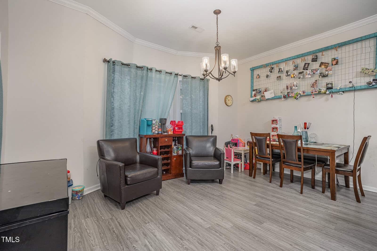 A room with two dark leather chairs, a small bookshelf, and a play kitchen under a window with blue curtains. A dining table with four chairs is against the wall, which features a large grid with photos and small items clipped to it.