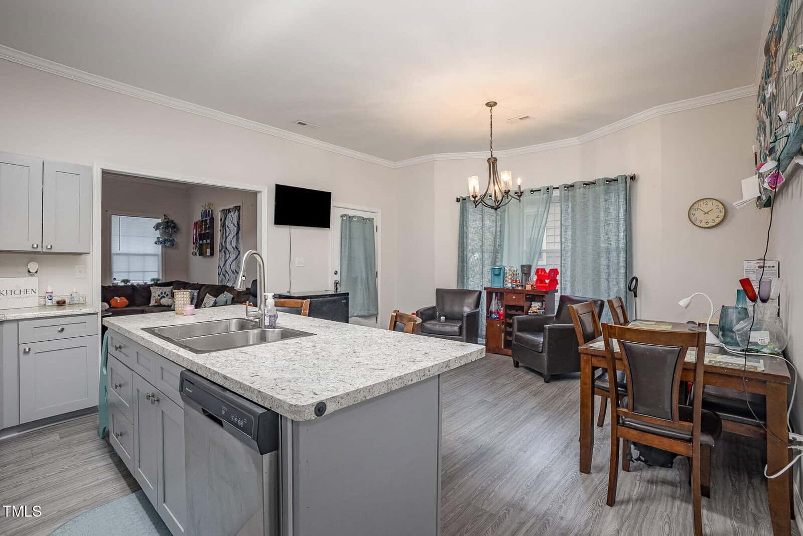 A modern kitchen and dining area with an island and stainless steel sink in the foreground. The dining space has a wooden table with chairs, a chandelier, and a seating area with armchairs. Artwork and a wall clock are visible on the walls.