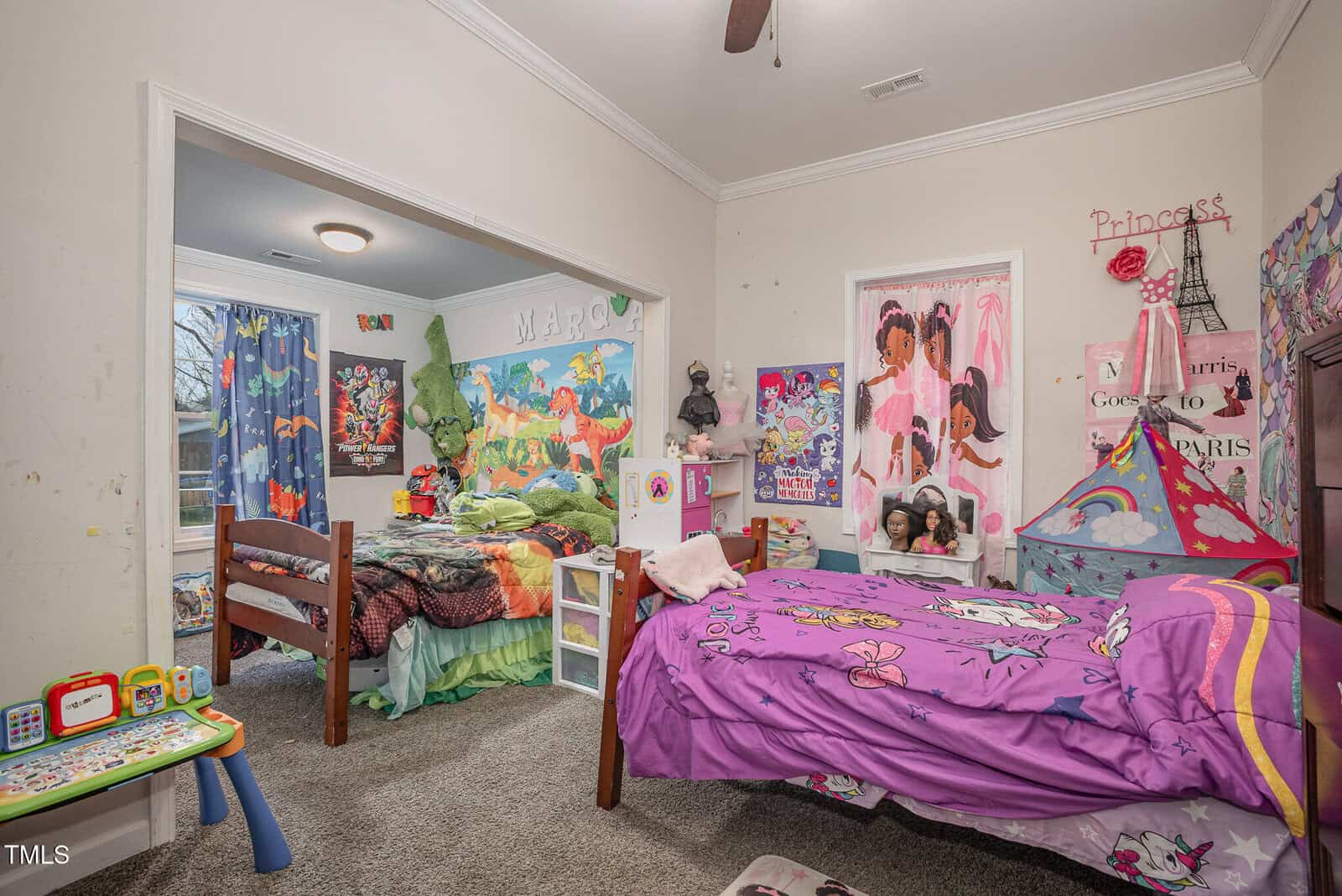 A children's bedroom with two beds. One bed is purple with a princess theme, while the other features green dinosaur bedding. The room has various wall decorations, including posters and artwork. A play tent is visible near the purple bed.