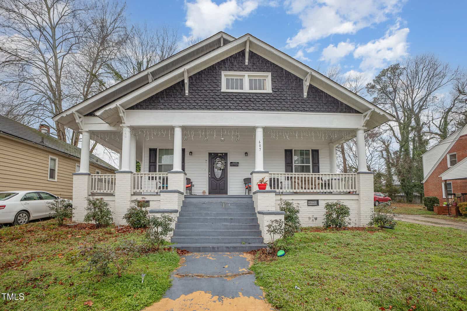 A two-story house with a dark shingle roof and white siding. The porch features white columns and railings, with steps leading to the front door. Two potted plants sit on either side of the entrance. A car is parked in the driveway beside the house.