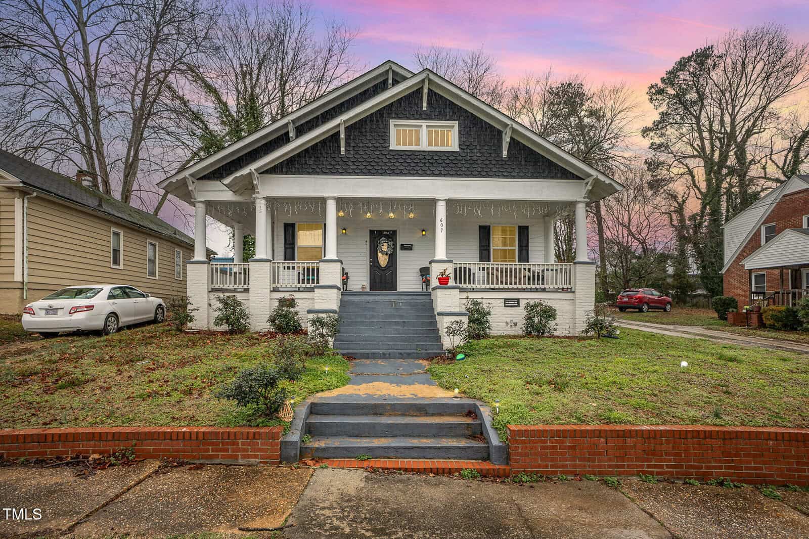 A charming two-story craftsman-style house at 607 Young Street, Henderson, boasts a large porch with steps leading up to it. The exterior features brick and siding, adorned with Christmas lights. A car rests in the driveway to the left under a sky painted in pink and purple hues at sunset.