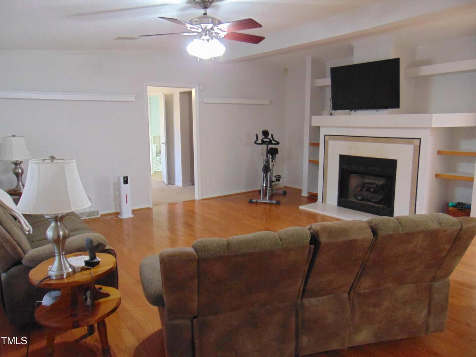 A cozy living room with brown sofas, wooden floors, a ceiling fan, a fireplace with a TV above it, an exercise bike, and shelves on either side of the fireplace. A side table with a lamp is next to the sofa.
