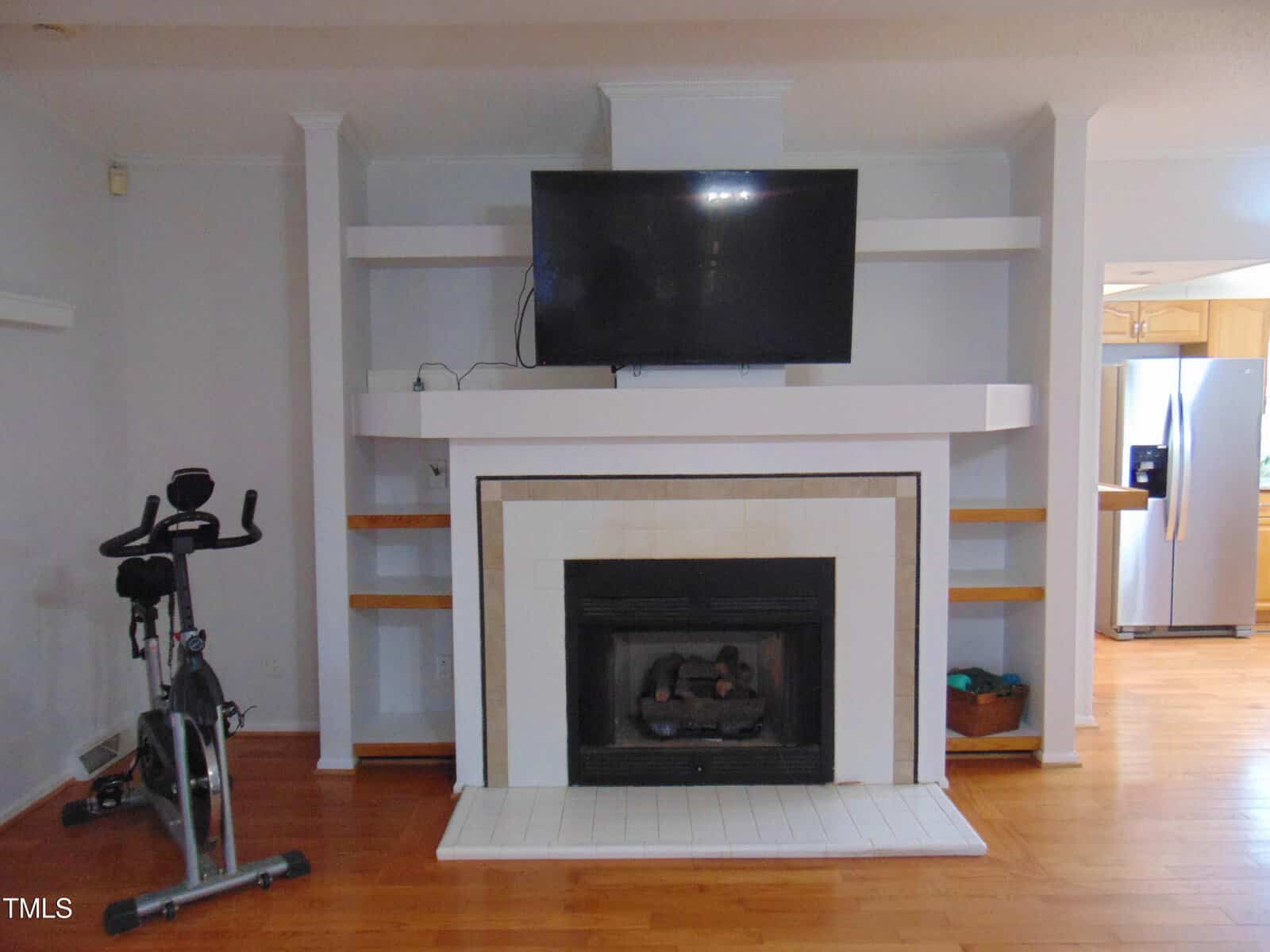 A living room with a mounted flat-screen TV above a white fireplace, built-in shelves on both sides, a stationary exercise bike to the left, and a view of a kitchen with a stainless steel fridge on the right.