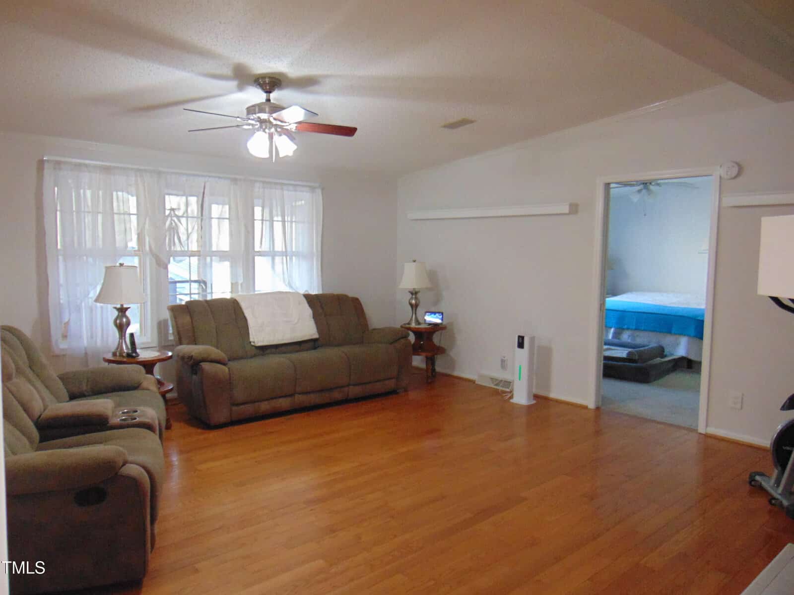 A living room with wooden floors, two brown sofas, two side tables with lamps, a ceiling fan with lights, and large windows with sheer curtains; a doorway leads to a bedroom with a visible bed.