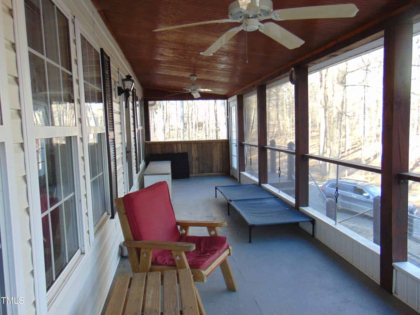 Screened-in porch with a wooden ceiling, ceiling fans, a red cushioned chair, a wooden table, and a lounge chair. Large windows line the left wall, and trees and a parked car are visible outside.