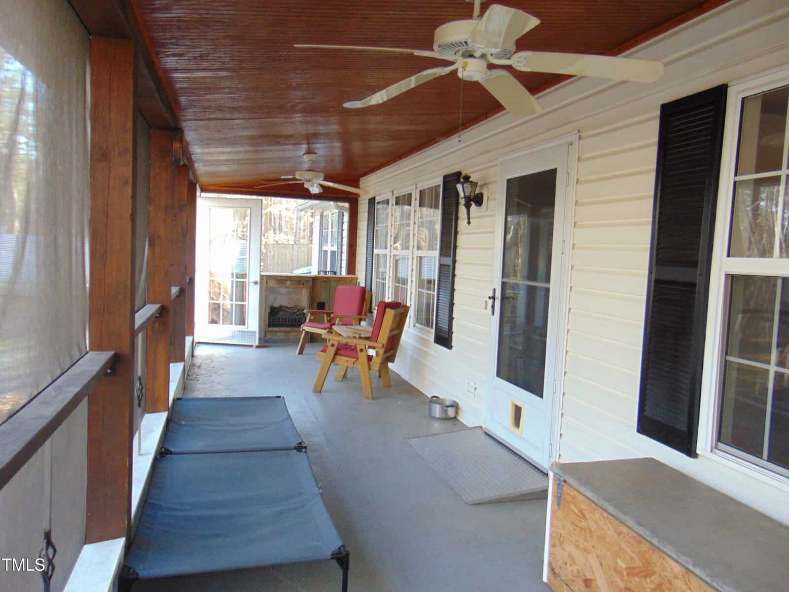 A screened-in porch with a wooden ceiling, ceiling fans, two red chairs and a small table, two pet loungers, and a white door and windows along the house wall. Bright natural light fills the space.