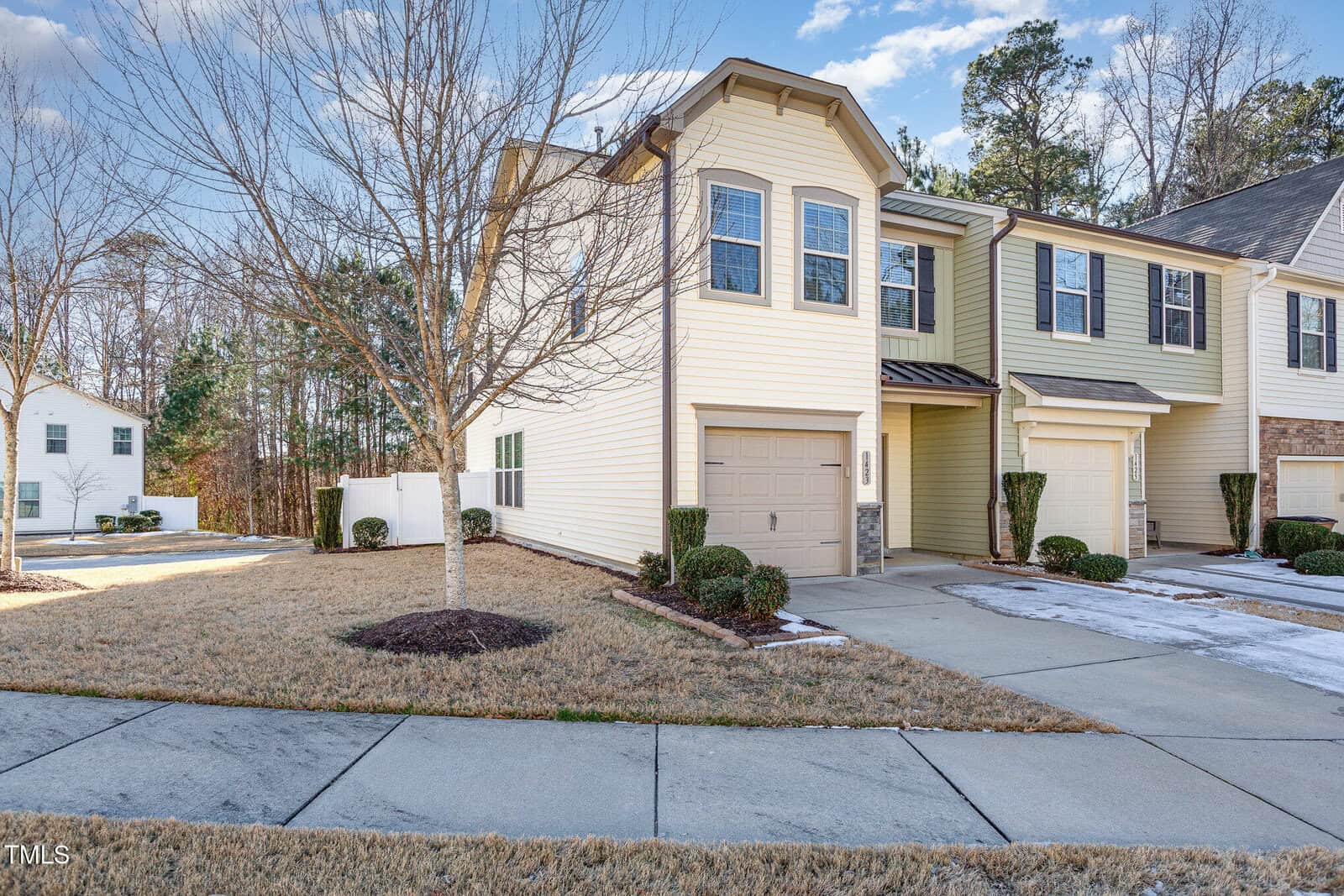 A two-story townhouse with beige siding and an attached single-car garage is pictured. The front yard has brown grass, a leafless tree, and trimmed shrubs. A concrete driveway and sidewalk are visible.