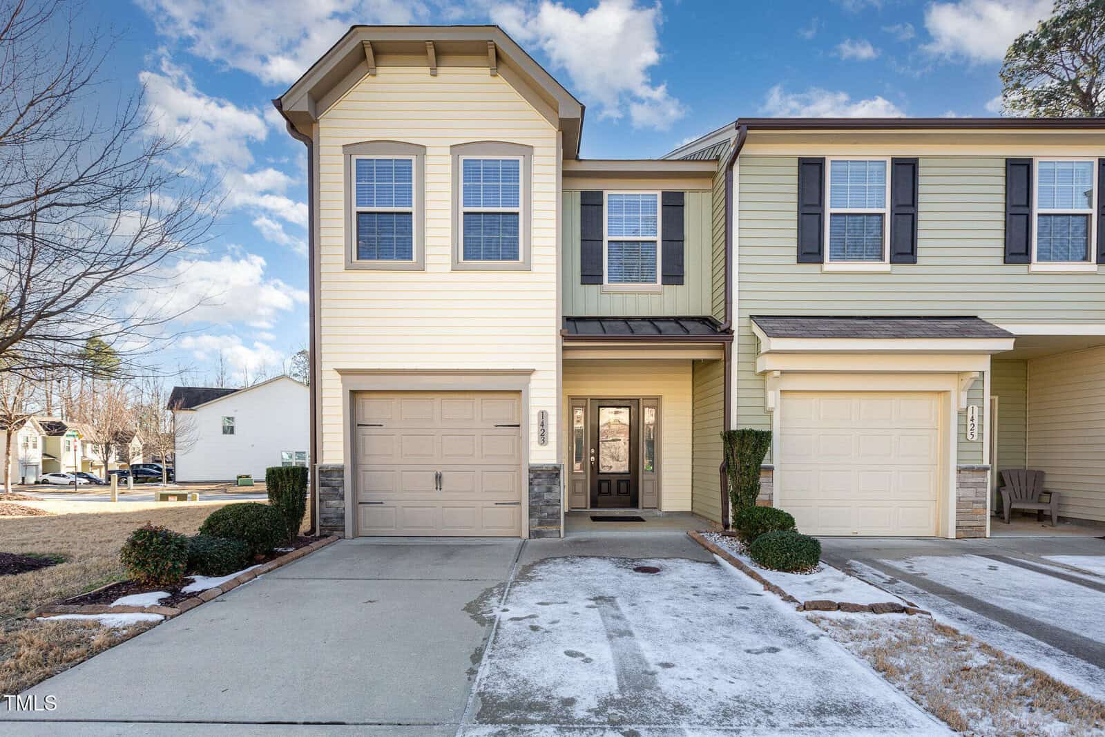 A two-story beige townhouse with a single-car garage, black shutters, and a small covered front entry. Patchy snow covers parts of the driveway and lawn. The sky is mostly clear with a few clouds.