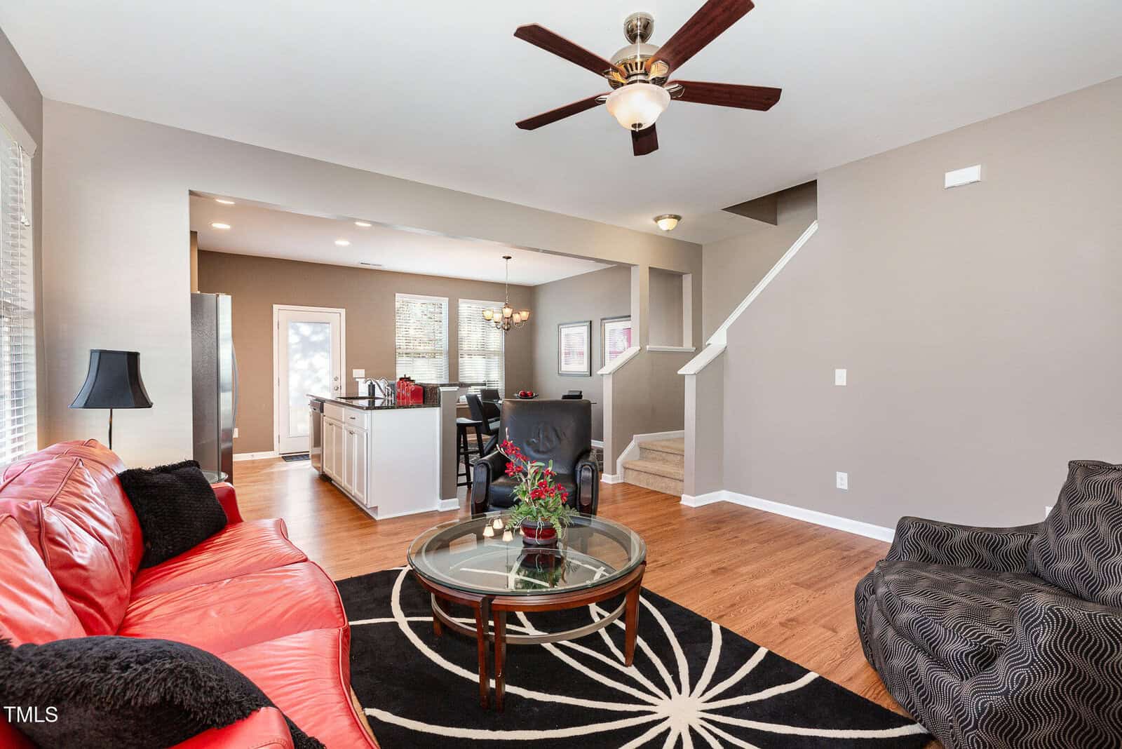 A living room with a red sofa, patterned armchair, glass coffee table, and black-and-white rug. The area opens to a kitchen and dining space. Stairs lead up, and a ceiling fan hangs from the white ceiling.