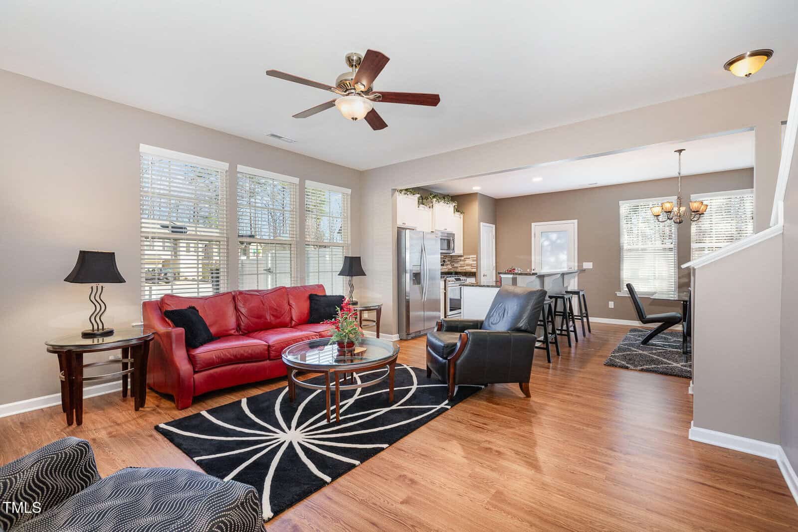 A modern open-concept living and dining area with wood flooring, a red sofa, black accent chairs, and round tables. A ceiling fan is above, and large windows let in natural light. The kitchen and dining area are visible in the background.