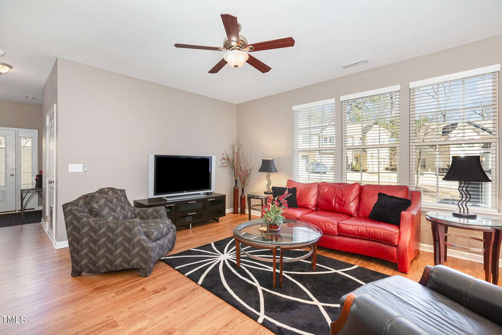 Bright living room with a red sofa, patterned armchair, TV on a stand, round glass coffee table, two lamps, and a large window with blinds letting in natural light. Wooden floors and neutral-colored walls.