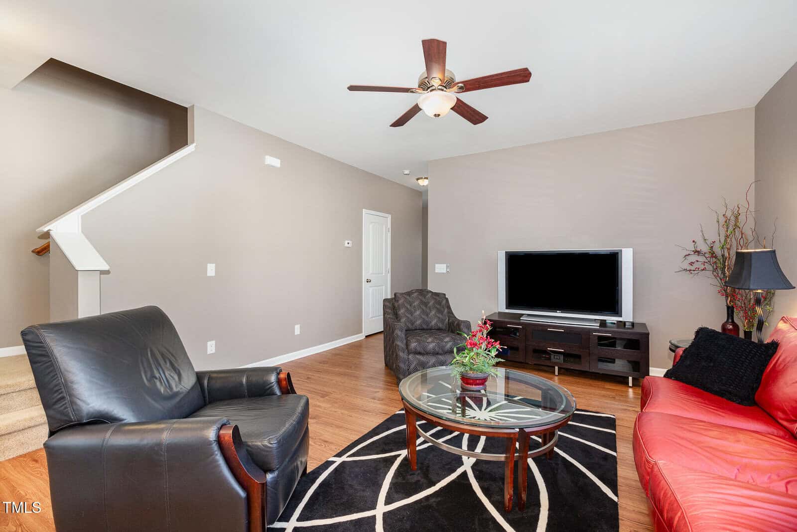 Modern living room with black armchair, red sofa, glass coffee table, TV on a stand, decorative plant, and wooden flooring. A staircase is visible to the left. Walls are light beige, and a ceiling fan is overhead.